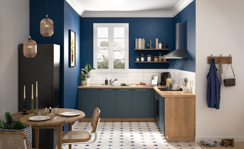 L-vormige keuken in Alby-blauw met werkblad en elementen in licht hout, wandplanken, en vloer met blauw gedessineerde cementtegels.